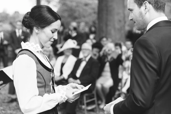 Black and white candid shot of the bride reading her vows from a folded card, her expression focused and emotional as the groom faces her. Intimate outdoor ceremony moment.