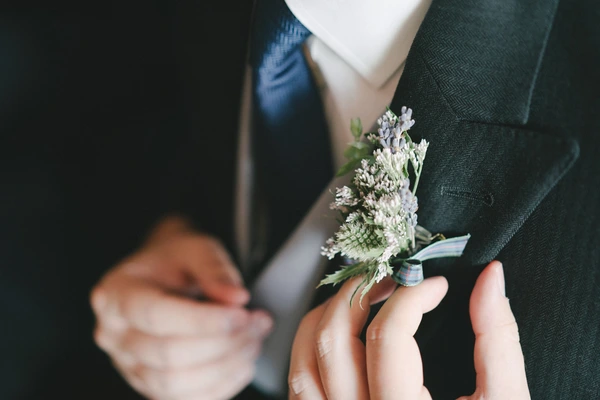 Groom in dark green suit holds a tiny white flower boutonniere, bride blurred in white behind