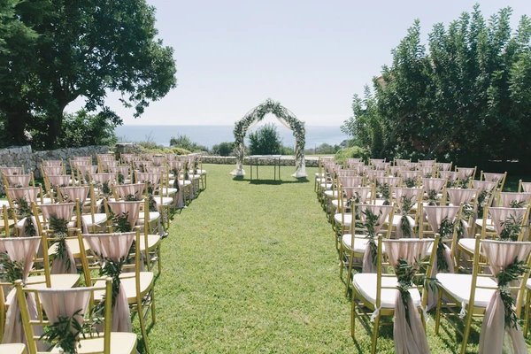 Outdoor ceremony on a green meadow — pink fabric draped chairs, green arch, sea view