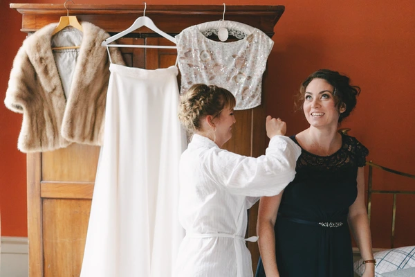 Two women in an orange room — one shows the white wedding dress to the other, who looks on with surprise and a smile.