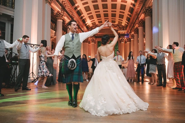 Groom in Scottish kilt and bride in white lace dance at the reception — guests close together, baroque hall with warm orange dome light.