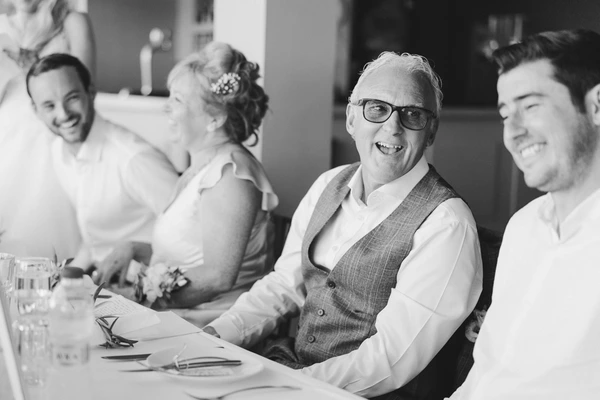 Older man in grey vest and white shirt smiles at the table, bride and young guests in background. B&W.