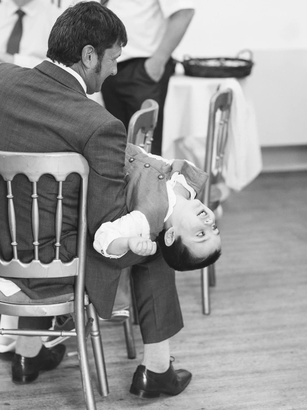 A man in a grey suit holds a toddler boy upside down playfully on a chair during a wedding reception. Black and white candid documentary moment.