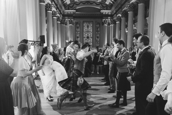 Bride and groom dancing in an imposing classical hall — laughing, guests cheering around them. Black-and-white.