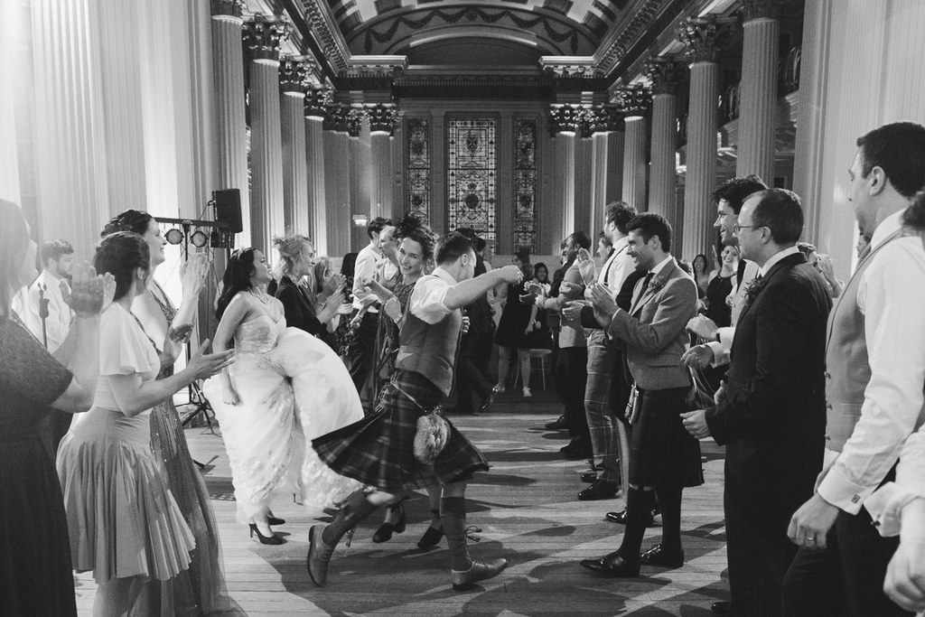 Bride and groom dancing in an imposing classical hall — laughing, guests cheering around them. Black-and-white.