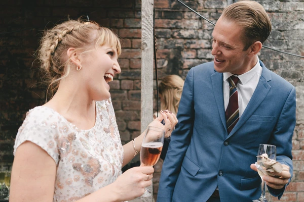 Bride and groom laughing and toasting at a brick venue — white lace, blue suit