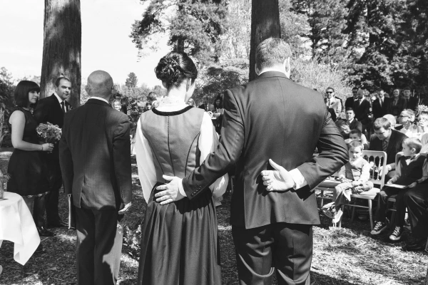 Black and white back view of the bride and her father arriving at the altar among tall trees, guests seated on either side. Candid processional documentary photo.