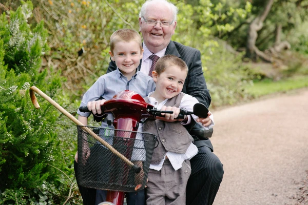 An older gentleman steadies two young boys sitting on a red bicycle on a garden path. Candid documentary wedding photography at the Royal Botanic Garden Edinburgh.