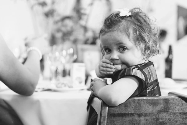 A toddler girl with a flower in her hair chews on food while leaning over a wedding reception chair, looking directly at the camera. Candid black and white documentary photograph.