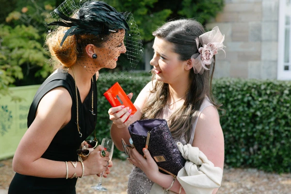 Two female guests in fascinator hats chatting animatedly at the outdoor reception, one in black with a feathered headpiece and the other in a pink floral dress. Candid drinks reception shot.