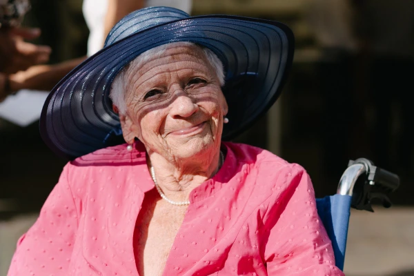 Elderly woman smiling wearing pink outfit and navy straw hat at outdoor garden wedding venue.