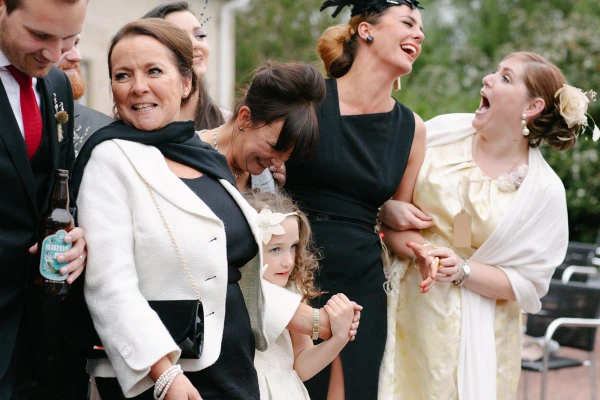 A candid group moment outside the wedding venue as guests of various ages react with laughter and delight, including a flower girl holding onto a woman's white jacket. Candid documentary wedding photography.