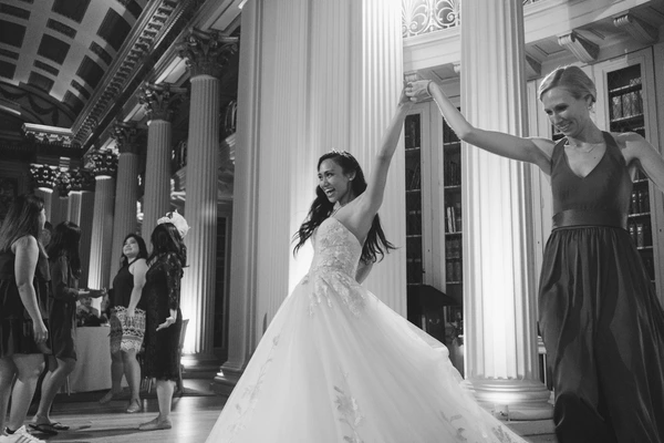 Bride in white lace twirls as a friend raises her arms to celebrate — white columns, happy moment