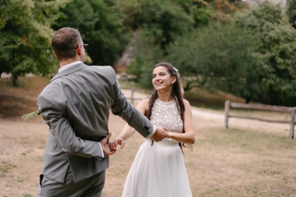 Bride and groom walking together holding hands at outdoor wedding with green trees and fence.