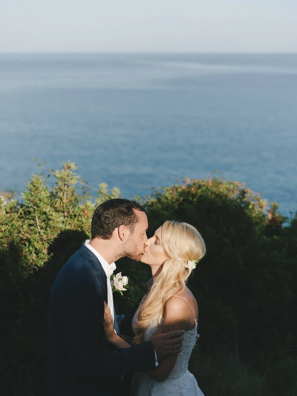 Bride and groom kiss on a cliff top above the sea — white lace, white flower hair pin, blue suit