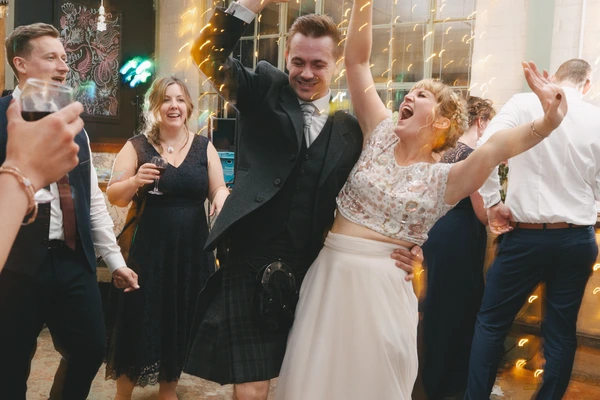 Bride raises her arms with joy on the dance floor, groom smiling beside her, guests celebrating