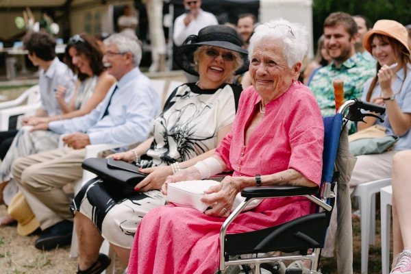 Elderly wedding guests smiling during outdoor ceremony, woman wearing pink hat and colorful scarf.