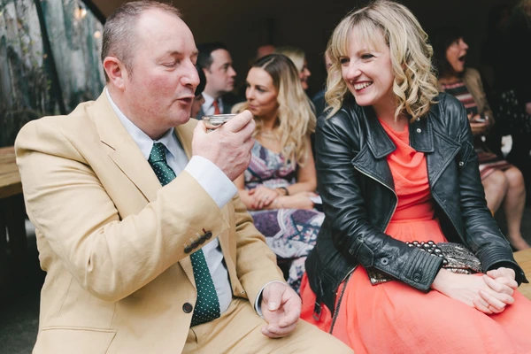 Man in cream suit and green tie, woman in red dress smiling — guests seated in background