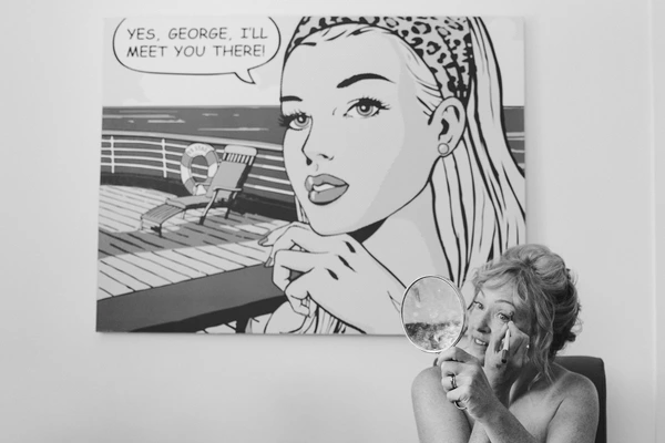 Woman laughs holding a mirror, looking at a pop art print — ring visible, playful black-and-white