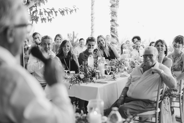 Wedding guests seated around the table in cheerful conversation — white tablecloth, flowers, palm trees
