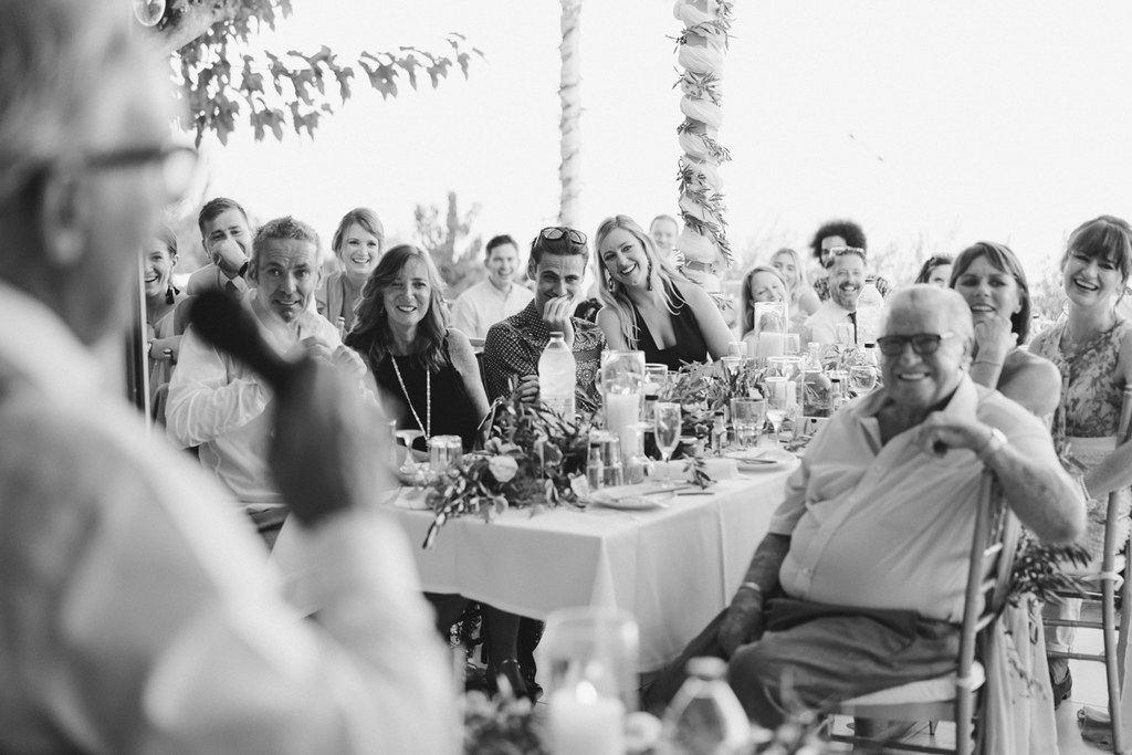 Wedding guests seated around the table in cheerful conversation — white tablecloth, flowers, palm trees