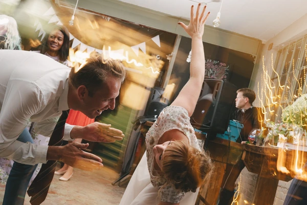 Bride in white lace raises her arms dancing at the wedding party, warm golden light and decor
