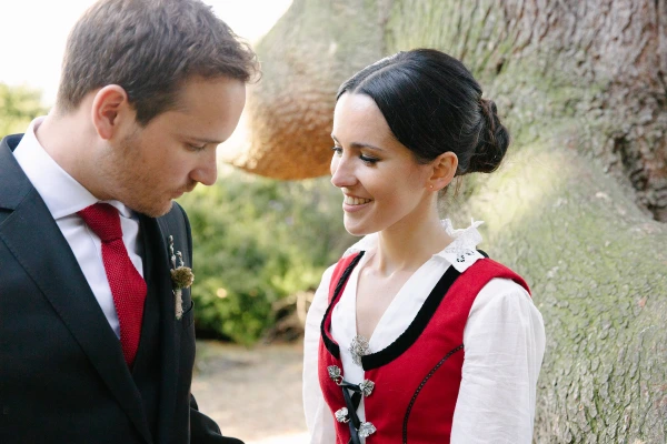 Bride in her red Norwegian bunad smiling warmly at the groom who looks down at her, both leaning against a large textured tree trunk in the Royal Botanic Garden Edinburgh. Intimate couple portrait.