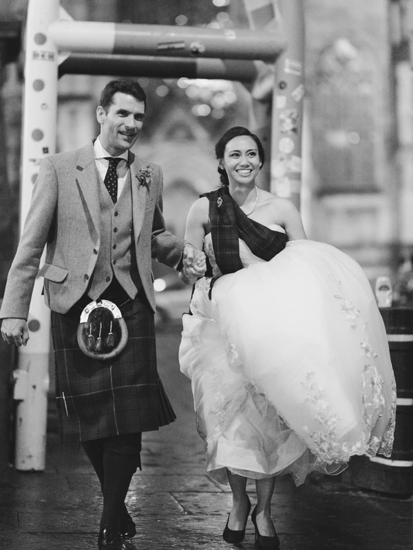 Bride and groom hold hands under the sweeping bridal veil before the venue, both smiling