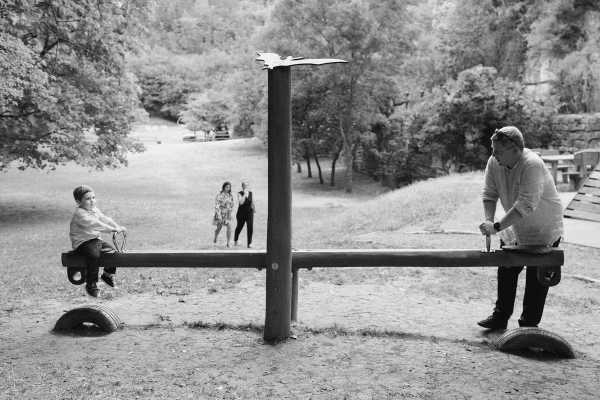 Black and white photo of two men playing seesaw at outdoor wedding with bride and guests watching.