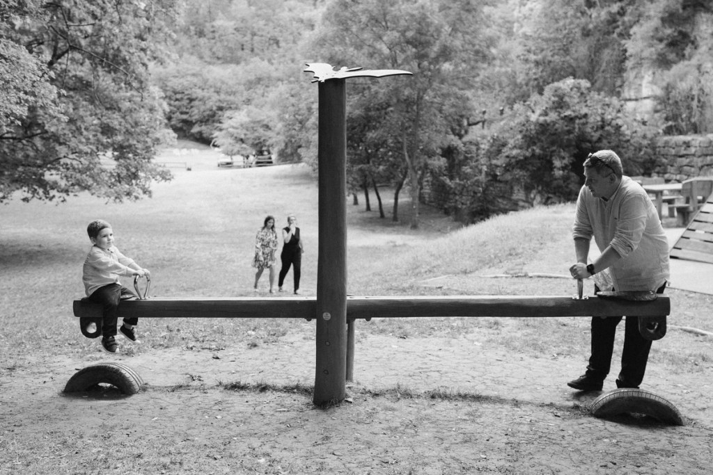 Black and white photo of two men playing seesaw at outdoor wedding with bride and guests watching.