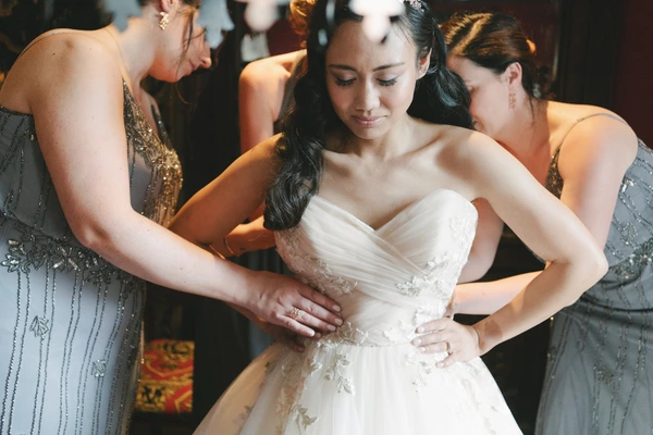 Bride in white stands as two women in black help with her gown — warm golden light, modest smile