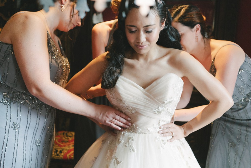 Bride in white stands as two women in black help with her gown — warm golden light, modest smile