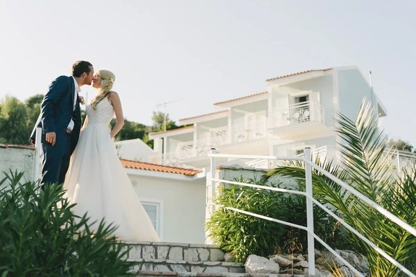 Bride and groom kiss on a rooftop terrace — palm trees and white balustrade, bright sunshine