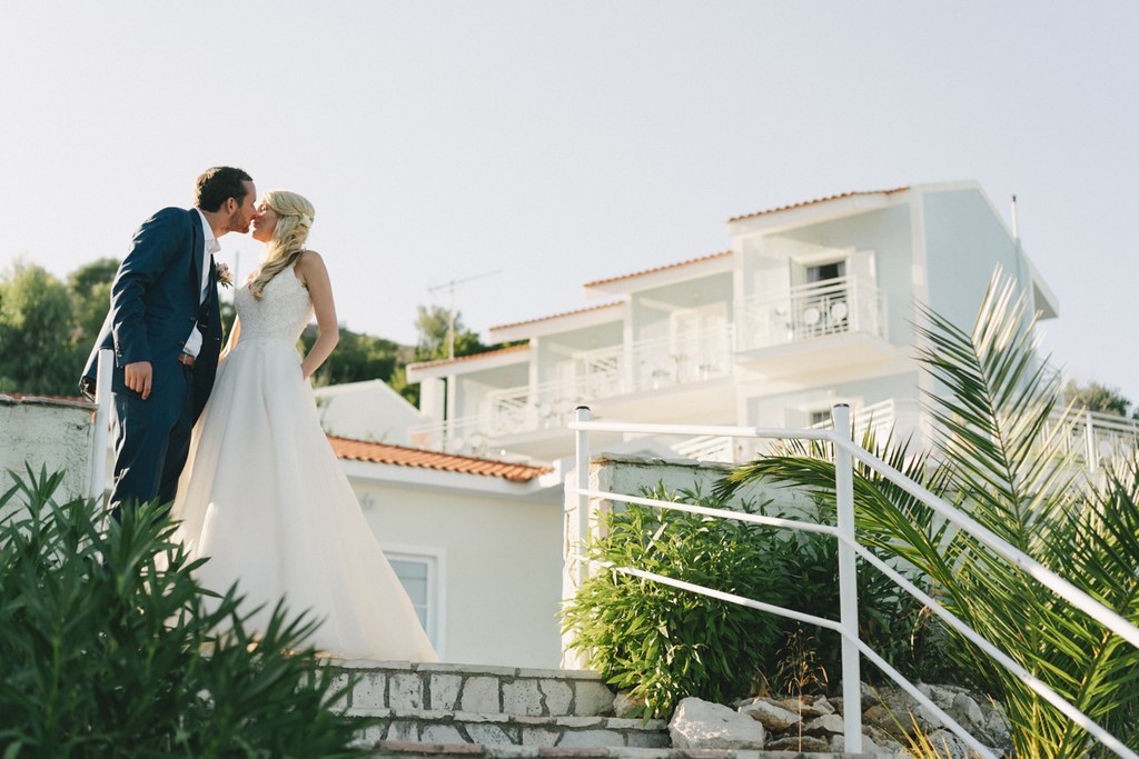 Bride and groom kiss on a rooftop terrace — palm trees and white balustrade, bright sunshine