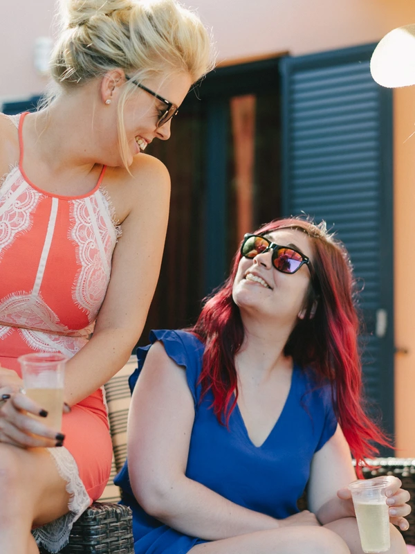 Two women laughing and talking at the wedding reception — red top, blue dress and sunglasses