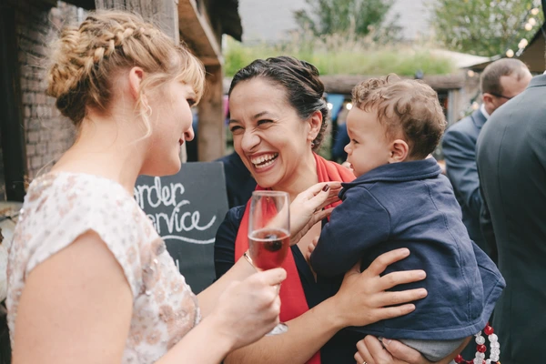 Woman in red scarf laughing while holding a small boy, bridesmaid with a souvenir glass beside her