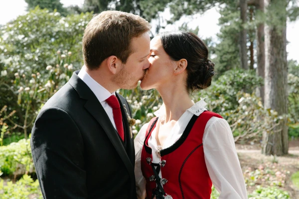 Couple kissing in the garden after the ceremony, bride in red Norwegian bunad and groom in a dark suit with red tie, surrounded by green trees. Joyful post-ceremony portrait.
