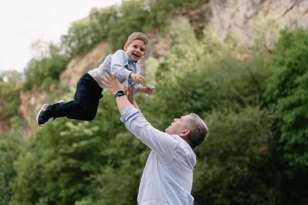 Man tossing young boy in air joyfully with rocky landscape and green trees in background.