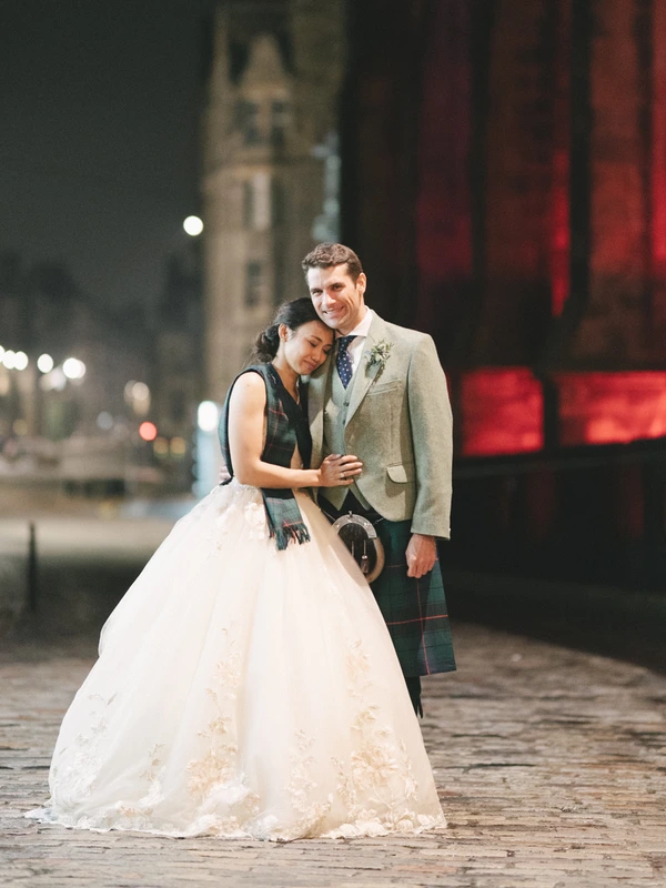 Night wedding portrait — bride in white lace rests in groom's arms, tartan kilt, red-lit city backdrop