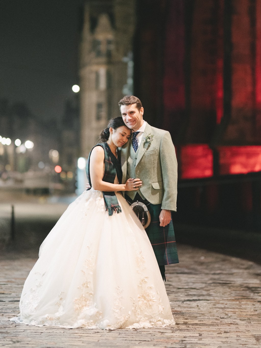 Night wedding portrait — bride in white lace rests in groom's arms, tartan kilt, red-lit city backdrop
