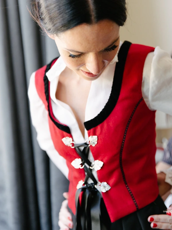 Bride in a red Norwegian bunad vest being helped with the lacing by a bridesmaid, while her father in a tuxedo with red bow tie watches from the doorway. Candid family moment.