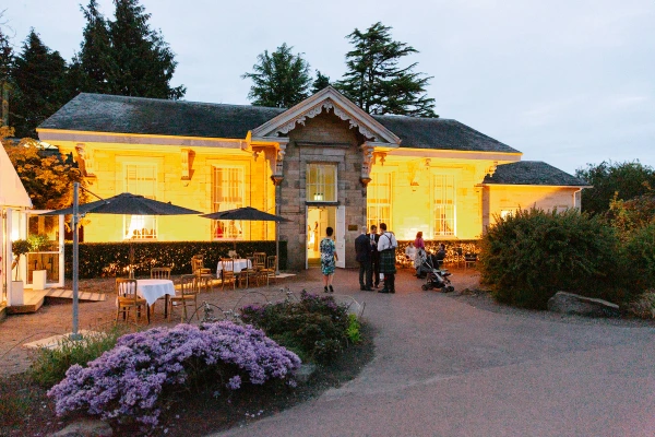 The wedding venue building at the Royal Botanic Garden Edinburgh glows warm gold at dusk, with a few guests visible on the terrace and flowering heather in the foreground. Documentary wedding photograph.