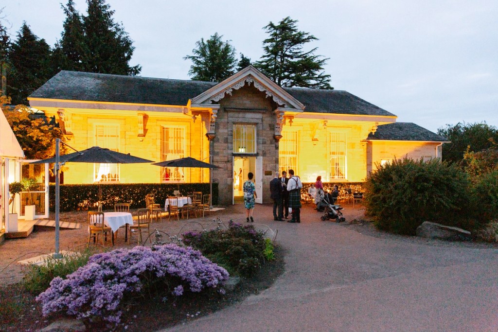 The wedding venue building at the Royal Botanic Garden Edinburgh glows warm gold at dusk, with a few guests visible on the terrace and flowering heather in the foreground. Documentary wedding photograph.