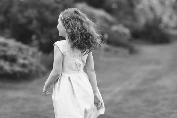 Black and white photograph of a flower girl in a white dress from behind, her curly hair caught by the wind as she walks across the garden lawn. Candid documentary wedding portrait.