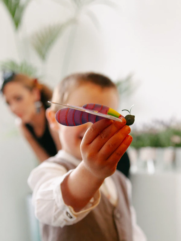 A toddler holds up a colourful toy butterfly close to the camera while a woman sits blurred in the background. Candid documentary wedding moment.