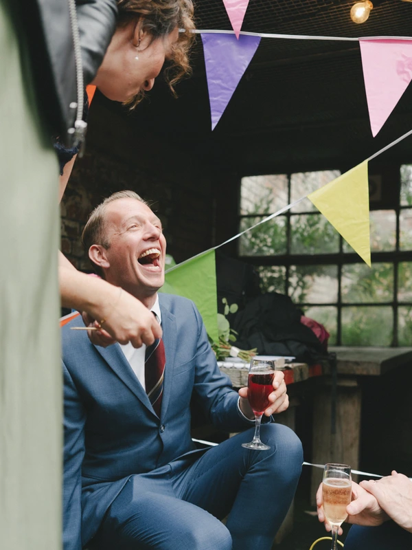 Groom in blue suit celebrating with a glass, a woman hugging him — colourful flags above