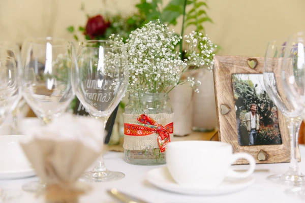 Wedding reception table detail with gypsophila in a hessian-wrapped jar, personalised wine glasses, and a framed family photograph. Rustic Scottish wedding decor.