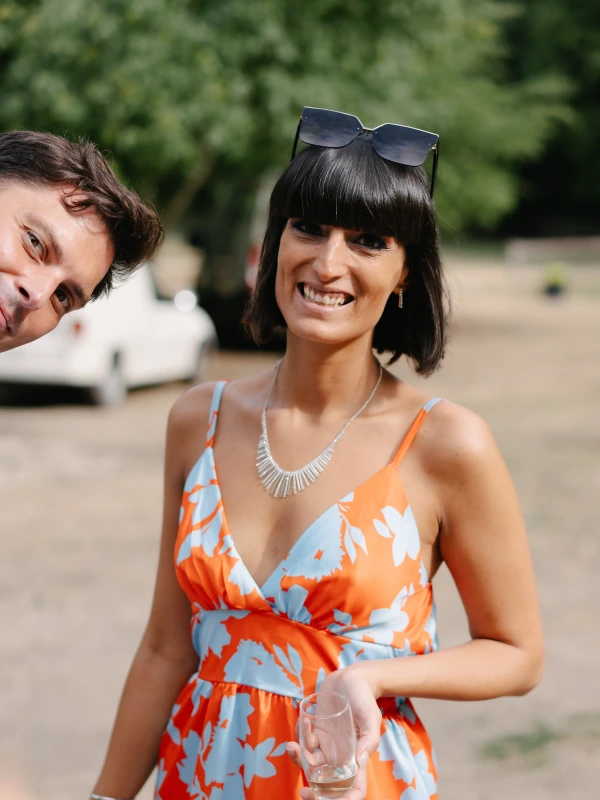 Woman in orange floral dress smiling at wedding reception with sunglasses and necklace accessory.