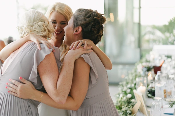 Bride smiling in embrace with two friends in grey bridesmaid dresses — floral centrepiece beside them