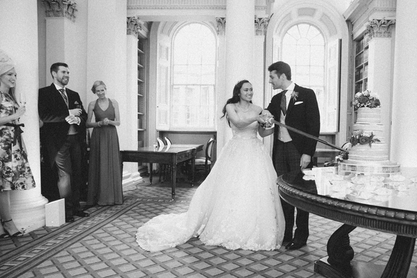 Bride and groom hold hands in the hall, guests watching — baroque interior, wedding cake on table